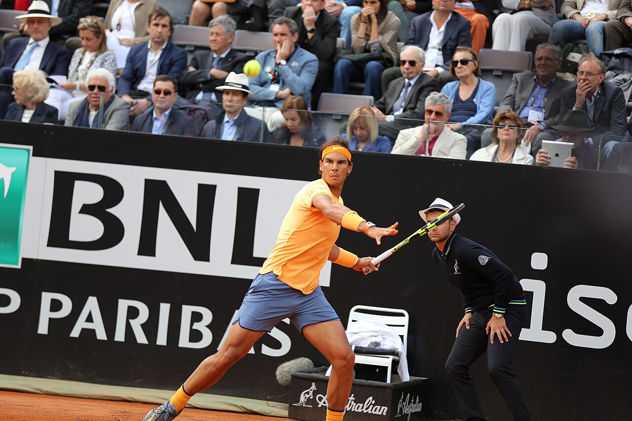 Speler in oranje shirt slaat een forehand op gravel